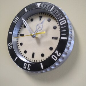 Puede incluir: Un reloj blanco y negro con un bisel plateado y una esfera blanca. El reloj tiene una aguja de segundos dorada y una aguja de minutos plateada. Los n&uacute;meros en el reloj son blancos y negros. El reloj est&aacute; colgado en una pared.
