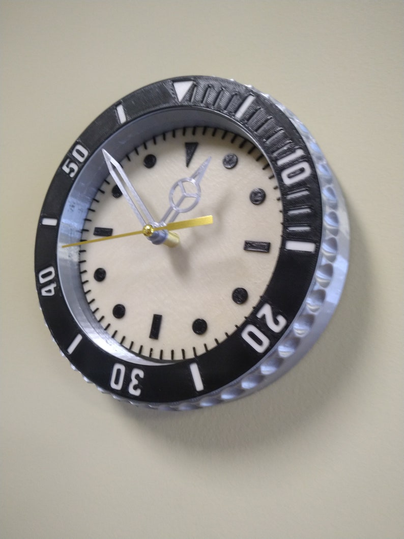 Puede incluir: Un reloj blanco y negro con un bisel plateado y una esfera blanca. El reloj tiene una aguja de segundos dorada y una aguja de minutos plateada. Los n&uacute;meros en el reloj son blancos y negros. El reloj est&aacute; colgado en una pared.