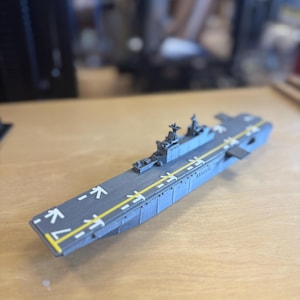 May include: A grey model aircraft carrier with yellow runway markings and directional arrows. The model has detailed structures on the deck and is set on a wooden surface.
