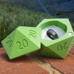 May include: A green 20-sided dice ring box with a silver ring inside. The box is open and the lid is laying next to it.