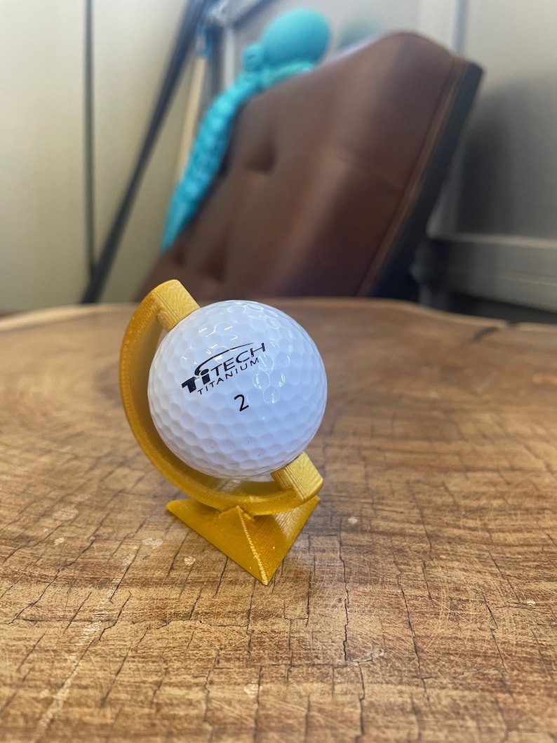 May include: A gold-colored golf ball holder with a white golf ball on top. The golf ball has the text "TiTech Titanium 2" printed on it.