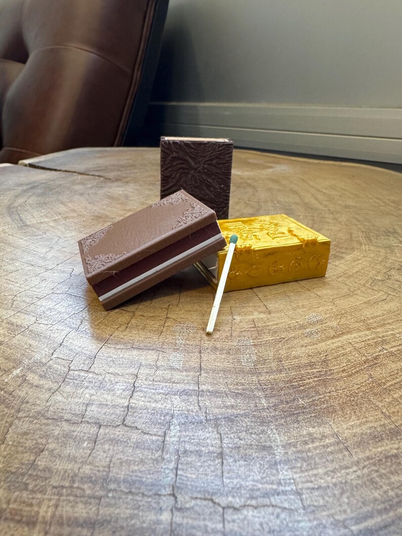 May include: Three small decorative boxes with intricate designs. The boxes are brown, gold, and a darker brown. A matchstick is laying on the table next to the boxes.