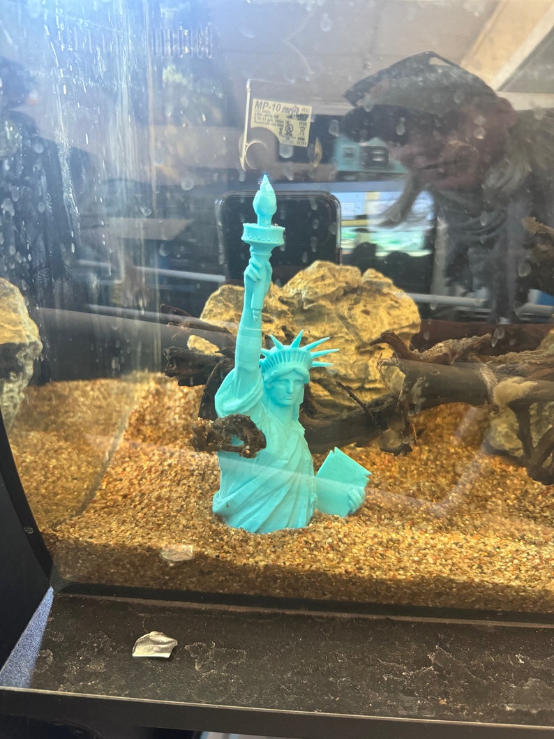K&ouml;nnte beinhalten: Eine t&uuml;rkisfarbene Plastikfigur der Freiheitsstatue in einem Aquarium mit braunem Kies und einem St&uuml;ck Treibholz.