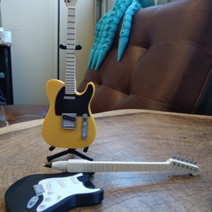 May include: Two miniature electric guitars, one yellow with a black pickguard and one black with a white pickguard, are displayed on a wooden surface. The yellow guitar is on a stand, while the black guitar is lying on its side.