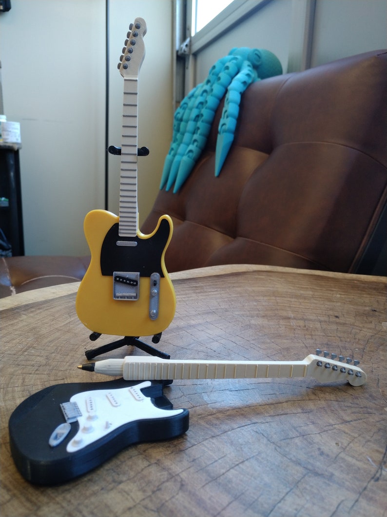May include: Two miniature electric guitars, one yellow with a black pickguard and one black with a white pickguard, are displayed on a wooden surface. The yellow guitar is on a stand, while the black guitar is lying on its side.