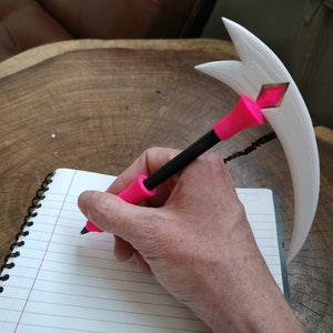 Puede incluir: Un portaplumas impreso en 3D de color blanco con forma de media luna con un bol&iacute;grafo rosa insertado. El bol&iacute;grafo est&aacute; siendo sostenido por una mano y est&aacute; escribiendo en un cuaderno.
