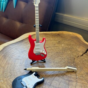 May include: Two miniature electric guitars, one red with a white pickguard and one black with a white pickguard. The guitars are on a wooden surface.