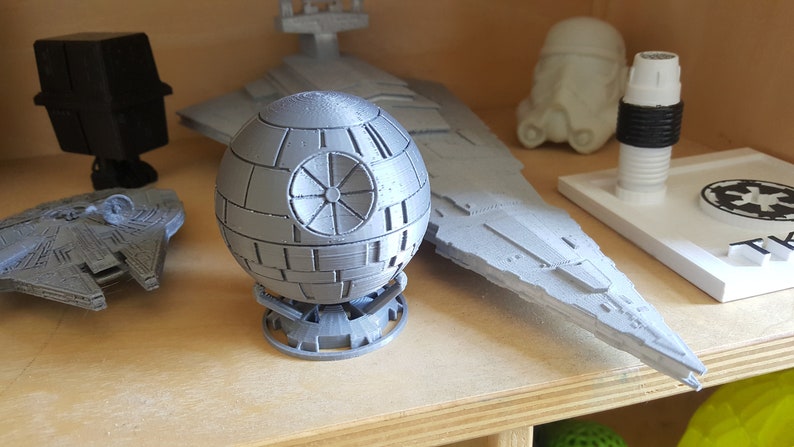 May include: A collection of gray 3D printed Star Wars models, including a Death Star, a TIE fighter, and a Star Destroyer. The models are displayed on a wooden shelf.