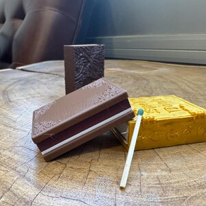 May include: Three small, decorative boxes with a vintage book design. The boxes are brown, yellow, and brown with a white stripe. A wooden matchstick with a green tip is lying on the table.