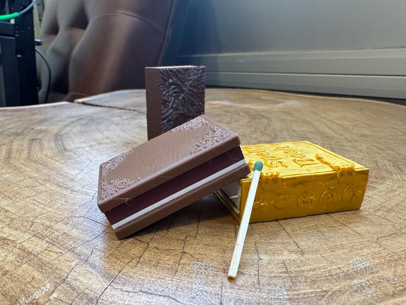 May include: Three small, decorative boxes with a vintage book design. The boxes are brown, yellow, and brown with a white stripe. A wooden matchstick with a green tip is lying on the table.