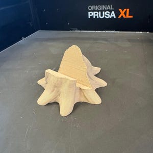 Puede incluir: Un objeto impreso en 3D en forma de estrella, de color marr&oacute;n claro, con una estructura central en forma de cono. El objeto est&aacute; hecho de material en capas y se encuentra sobre una superficie gris oscuro. El texto "ORIGINAL PRUSA XL" es visible en el fondo.