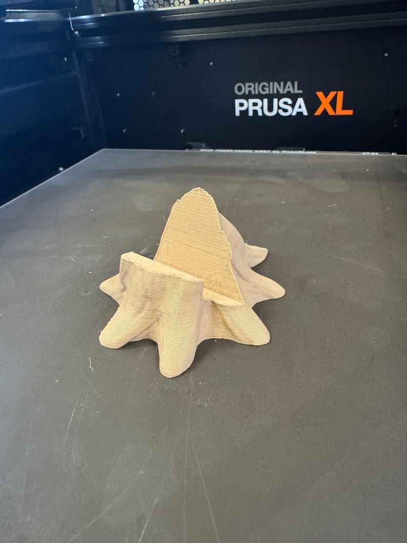 Puede incluir: Un objeto impreso en 3D en forma de estrella, de color marr&oacute;n claro, con una estructura central en forma de cono. El objeto est&aacute; hecho de material en capas y se encuentra sobre una superficie gris oscuro. El texto "ORIGINAL PRUSA XL" es visible en el fondo.