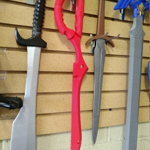 May include: Four different swords hanging on a wall. The swords are made of plastic and are painted in different colors. The swords are hanging on a wooden wall with a brick wall behind it.