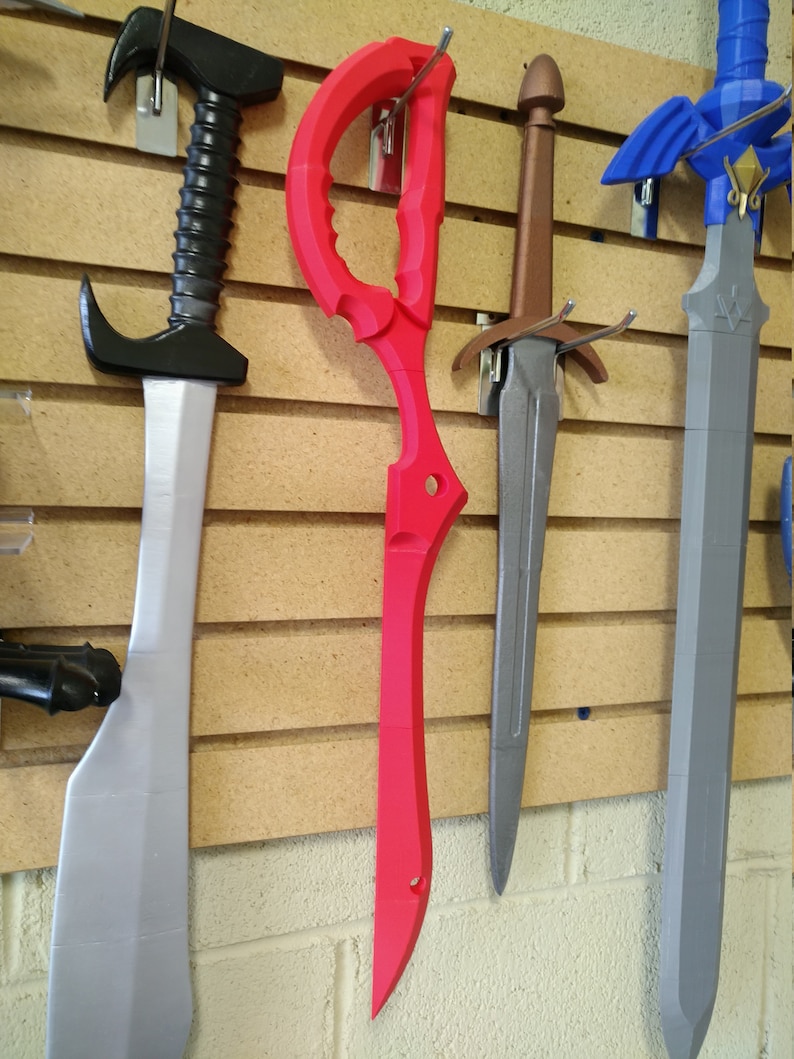 May include: Four different swords hanging on a wall. The swords are made of plastic and are painted in different colors. The swords are hanging on a wooden wall with a brick wall behind it.