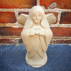 May include: A beige angel statue with folded hands and feathered wings. The statue has a serene expression and is set against a red brick wall and a dark blue surface. The angel's robe is detailed with soft folds.