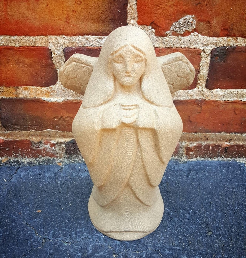May include: A beige angel statue with folded hands and feathered wings. The statue has a serene expression and is set against a red brick wall and a dark blue surface. The angel's robe is detailed with soft folds.