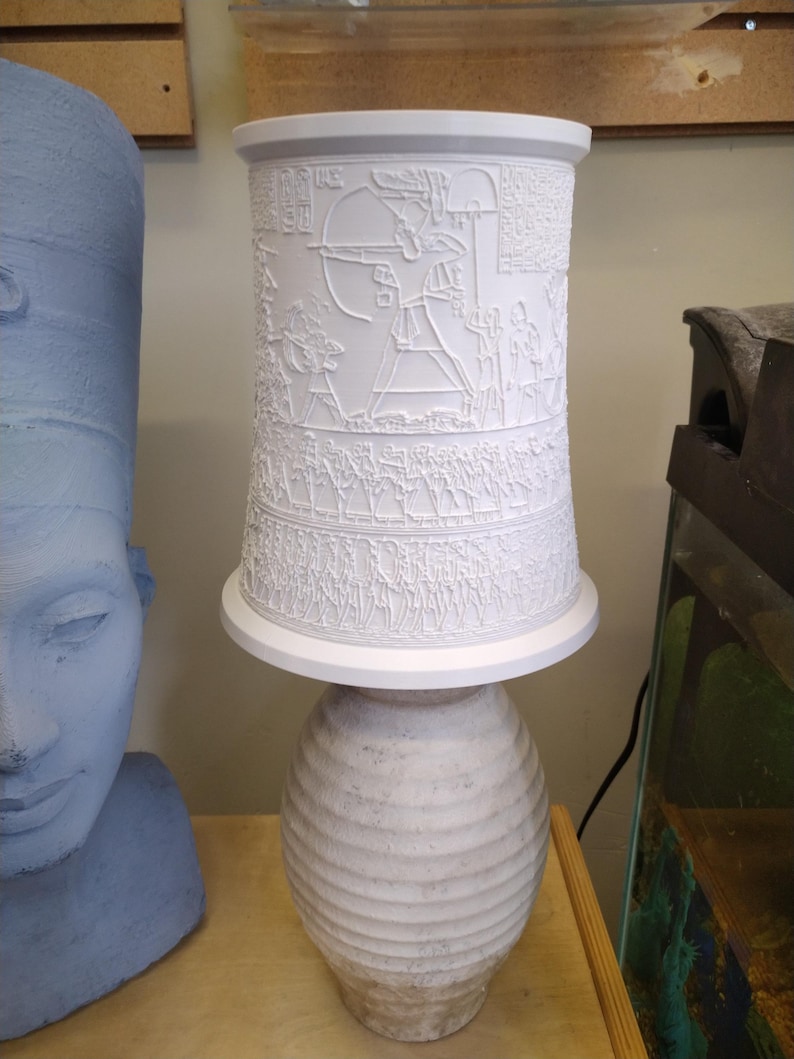 Pu&ograve; includere: Lampada in ceramica bianca con un paralume cilindrico che presenta un design in rilievo di geroglifici e una figura che tiene un arco e una freccia. La base della lampada &egrave; un vaso in ceramica bianca con un design testurizzato e a coste.