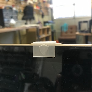 May include: A white plastic webcam cover with a shield design, attached to a laptop screen.