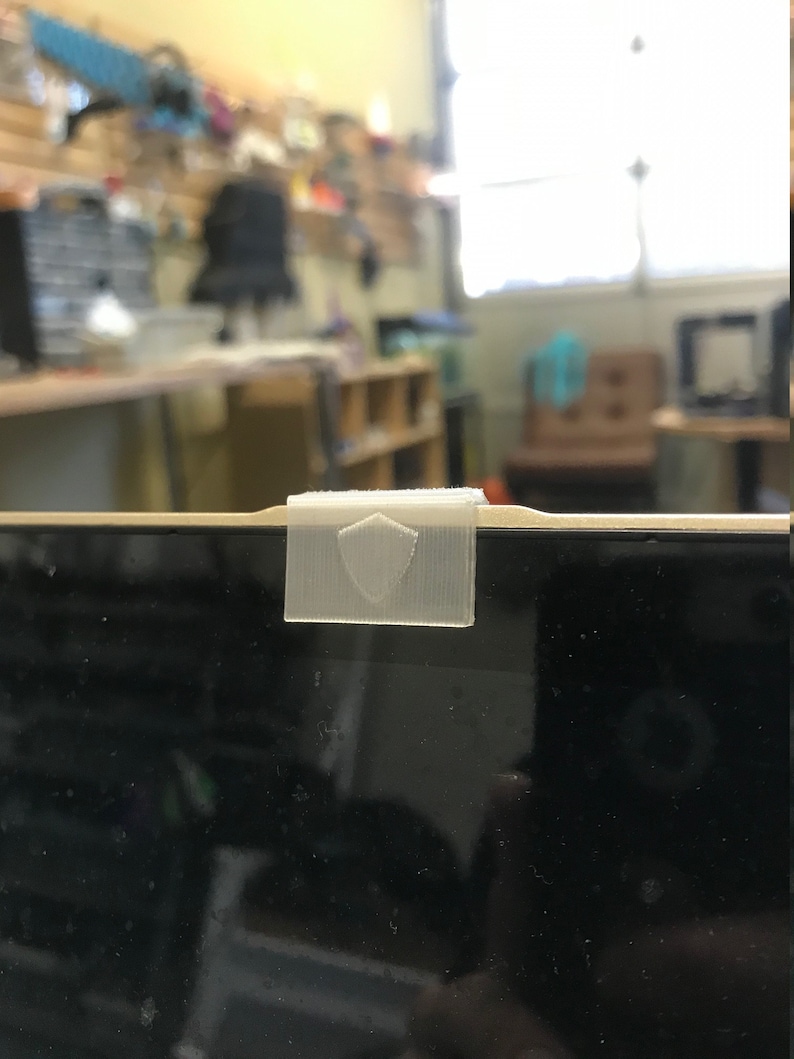 May include: A white plastic webcam cover with a shield design, attached to a laptop screen.