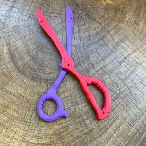 May include: A pair of scissors with one purple blade and one red blade. The scissors are lying on a wooden surface.