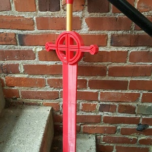 Op de afbeelding: Een rood plastic zwaard met een gouden handvat en een cirkelvormig ontwerp op de beschermkap. Het zwaard leunt tegen een bakstenen muur.
