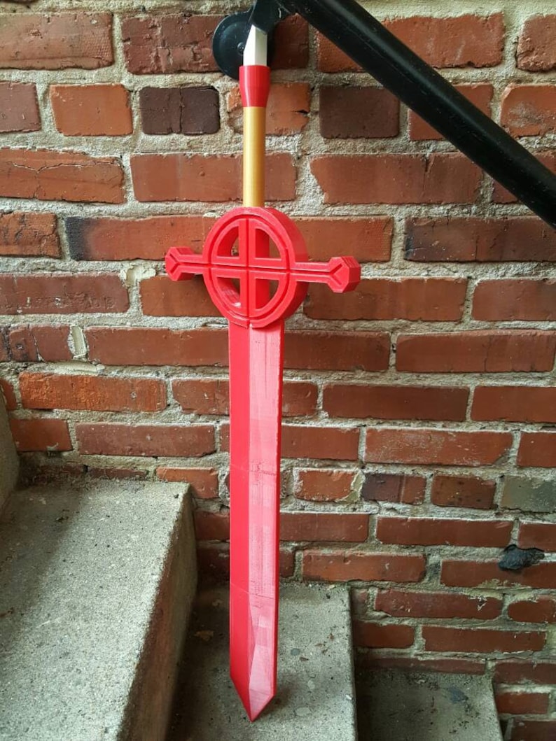 Op de afbeelding: Een rood plastic zwaard met een gouden handvat en een cirkelvormig ontwerp op de beschermkap. Het zwaard leunt tegen een bakstenen muur.