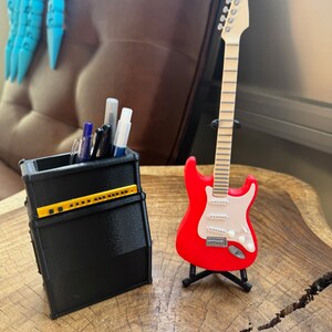May include: A black and yellow guitar amplifier-shaped pencil holder with pens and markers inside, next to a red electric guitar on a stand. The guitar has a white pickguard and a wooden neck.