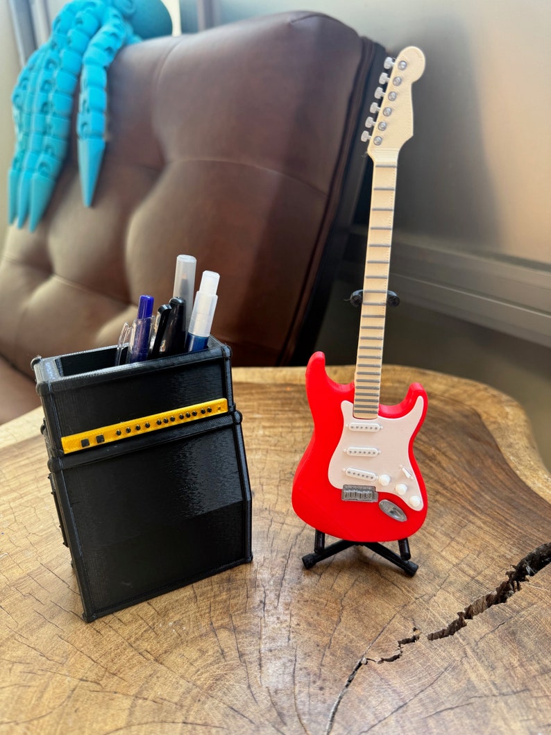 May include: A black and yellow guitar amplifier-shaped pencil holder with pens and markers inside, next to a red electric guitar on a stand. The guitar has a white pickguard and a wooden neck.