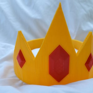 May include: A yellow plastic crown with three red gem accents. The crown is designed to be worn as a headband.