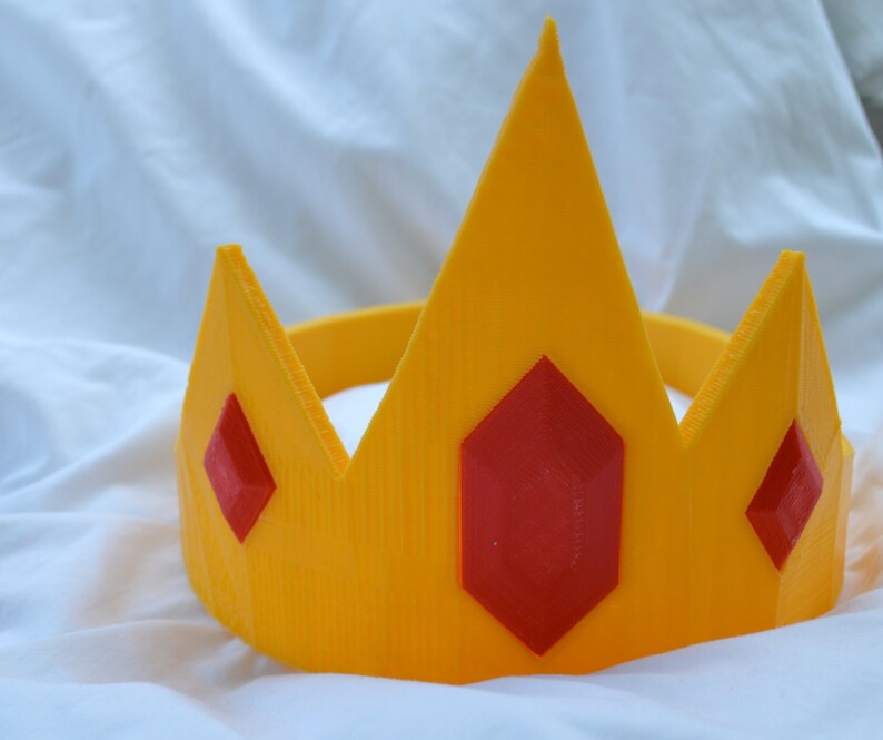 May include: A yellow plastic crown with three red gem accents. The crown is designed to be worn as a headband.