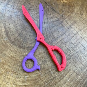 May include: A pair of scissors with one purple blade and one red blade. The scissors are lying on a wooden surface.