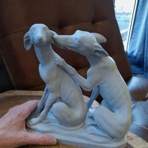 May include: A gray ceramic sculpture of two dogs, one sitting and the other standing, with their heads touching. The sculpture is on a wooden surface.
