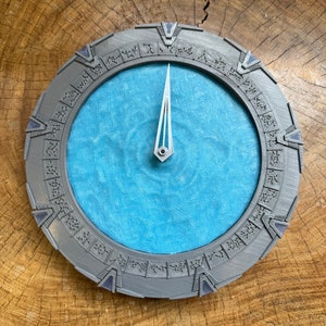 May include: A grey and blue circular clock with a silver hand pointing to the 12. The clock face is surrounded by a ring of symbols.