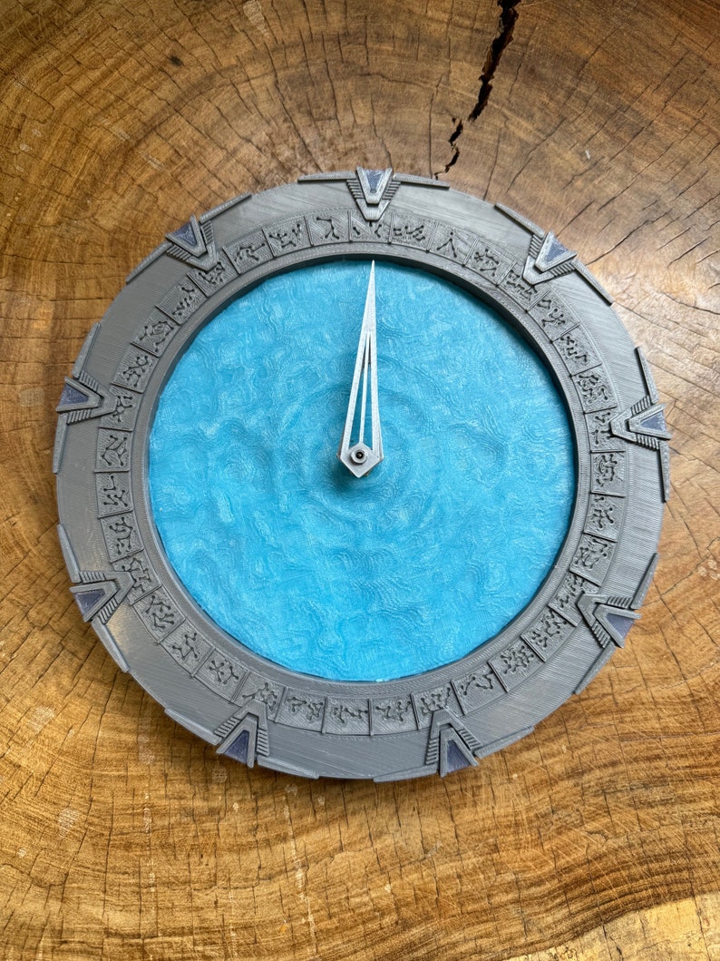 May include: A grey and blue circular clock with a silver hand pointing to the 12. The clock face is surrounded by a ring of symbols.