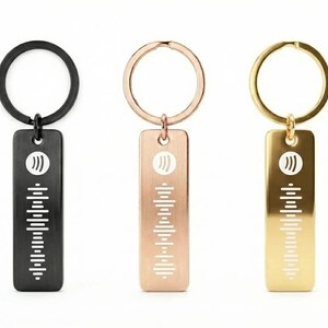 May include: Three keyrings in black, rose gold, and gold. Each keyring features a rectangular tag with a white sound wave design and a circular ring. The keyrings are shown against a white background.