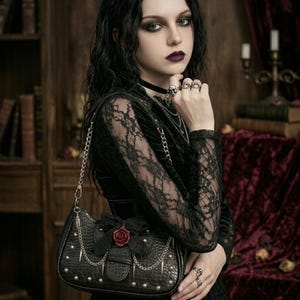 May include: Black handbag with a silver chain strap, studded details, and a red rose accent. The bag has a bow and spike details. The woman is wearing a black lace top and multiple rings.