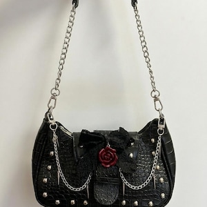 May include: Black shoulder bag with a crocodile texture, silver chain straps, and studded accents. The bag features a black lace bow with a red rose embellishment and dangling chain details, creating a gothic aesthetic.