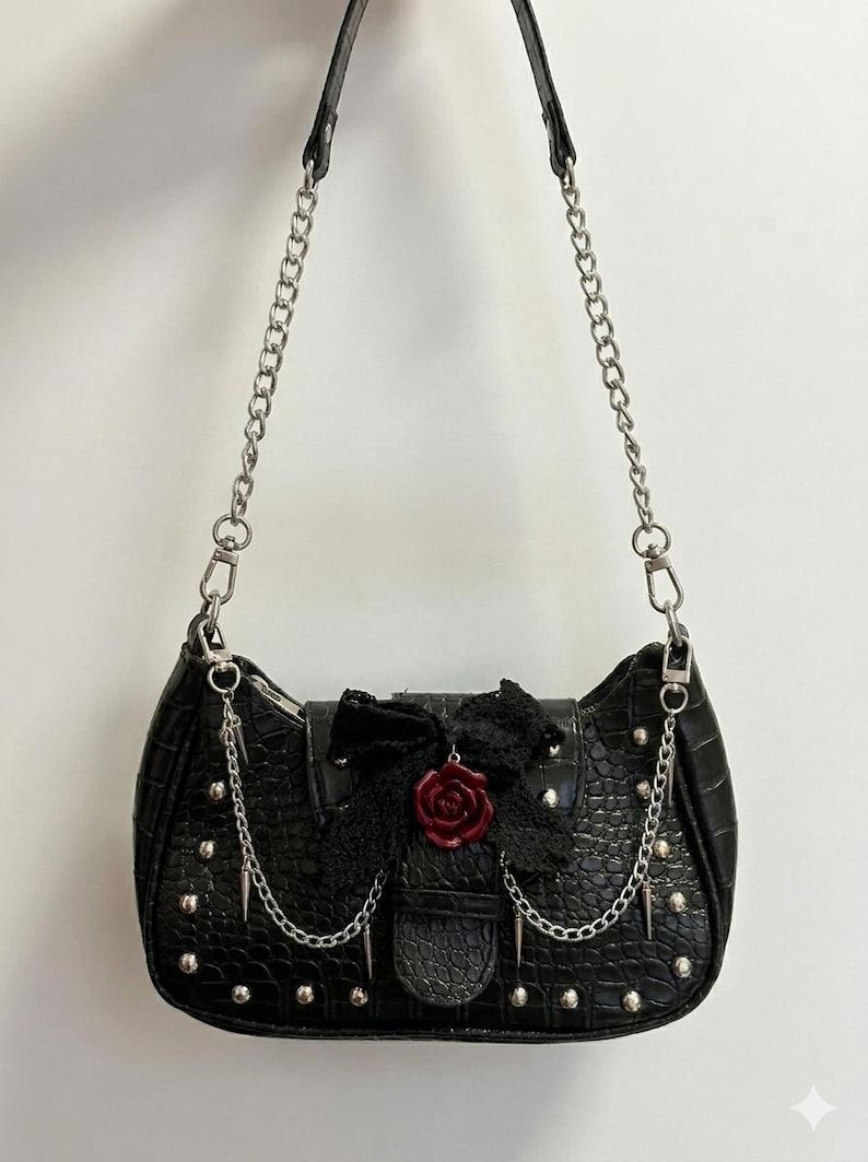 May include: Black shoulder bag with a crocodile texture, silver chain straps, and studded accents. The bag features a black lace bow with a red rose embellishment and dangling chain details, creating a gothic aesthetic.