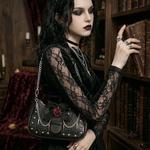 May include: Black handbag with a chain strap, studded details, and a red rose accent. The bag is part of a gothic-themed ensemble, with a black lace top and multiple silver necklaces. The image is set in a library with antique books.