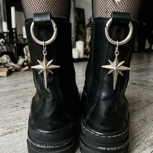 Può includere: Un paio di stivali in pelle nera con una spessa suola in gomma e la scritta FASHION IS FOR BRAVE stampata sul lato. Ogni stivale presenta un anello in metallo argentato fissato alla linguetta posteriore, da cui pende un ciondolo a stella.