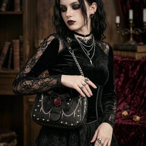 May include: Black handbag with silver chain straps, studs, and a red rose detail. The bag is held by a person wearing a black velvet top with lace sleeves, and multiple silver necklaces and rings. The style is gothic.