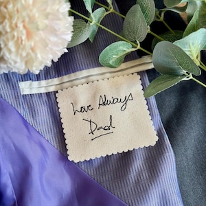Può includere: Una toppa quadrata in tessuto color bianco sporco con bordi smerlati che presenta il testo ricamato in nero Love Always Dad. La toppa è fissata all'interno di una giacca blu scuro con fodera a righe azzurre e una tasca in raso viola.