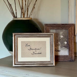 May include: A silver-framed print with the handwritten message "Love, Grandma & Grandad". The frame has a textured finish. A second framed picture and a dark green vase with dried plants are visible in the background.