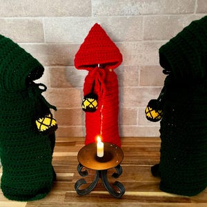 Pode incluir: Três capas de garrafa de crochê em verde e vermelho, que se assemelham a figuras com capuz. Cada figura segura uma pequena lanterna amarela. Uma vela acesa está em um suporte de metal decorativo, criando uma cena mística.