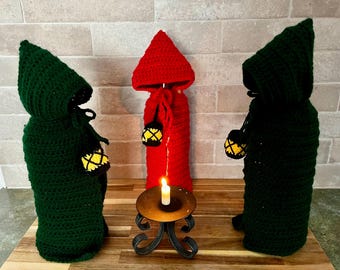 May include: Three crocheted bottle covers in green and red, resembling hooded figures. Each figure holds a small, yellow lantern. A lit candle sits on a decorative metal stand in front of the figures, creating a mystical scene.