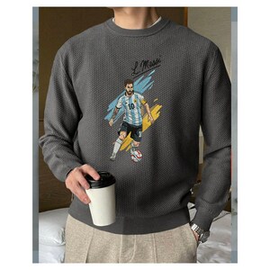 May include: A charcoal gray long-sleeve crewneck sweater with a textured waffle-knit pattern. The front features a graphic illustration of a soccer player in a light blue and white striped jersey with the number 10, accompanied by the text L. Messi.