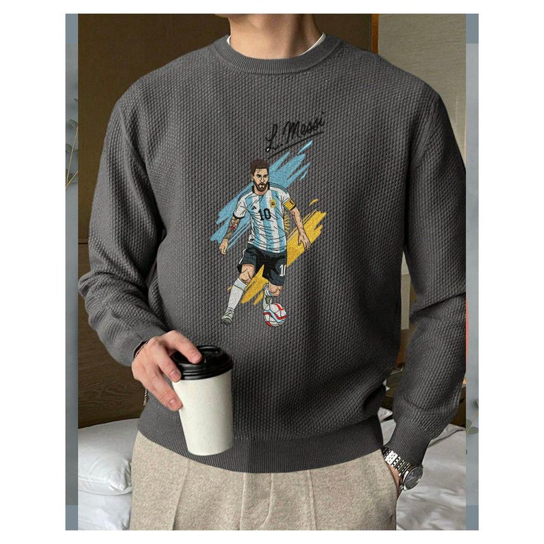 May include: A charcoal gray long-sleeve crewneck sweater with a textured waffle-knit pattern. The front features a graphic illustration of a soccer player in a light blue and white striped jersey with the number 10, accompanied by the text L. Messi.