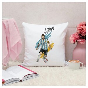 May include: A white square throw pillow featuring a graphic illustration of a soccer player wearing a light blue and white striped jersey with the number 10, black shorts, and white socks, dribbling a soccer ball, with the text L. Messi above.
