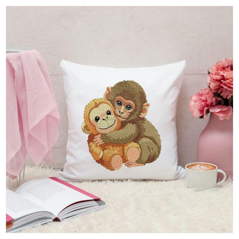 May include: White square pillow featuring an embroidered design of two hugging monkeys. One monkey is brown, and the other is tan. The pillow is on a white fluffy surface, with a pink vase and a cup of coffee nearby.