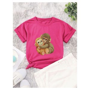 May include: A magenta t-shirt featuring a graphic of two hugging monkeys. The short-sleeved shirt is paired with white shorts and white sneakers. The shirt has a knotted detail at the waist.